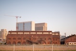 A large brick industrial building is seen in the foreground with multiple arched windows. Behind it are modern high-rise buildings with glass facades and a construction crane. The area around the industrial building is empty, with grass and a simple fence in the forefront.