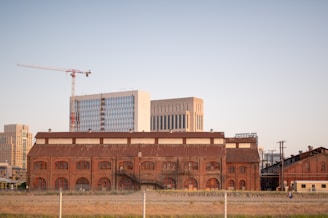 A large brick industrial building is seen in the foreground with multiple arched windows. Behind it are modern high-rise buildings with glass facades and a construction crane. The area around the industrial building is empty, with grass and a simple fence in the forefront.