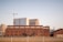 A large brick industrial building is seen in the foreground with multiple arched windows. Behind it are modern high-rise buildings with glass facades and a construction crane. The area around the industrial building is empty, with grass and a simple fence in the forefront.