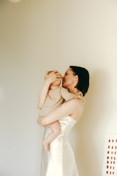 A woman is holding a child in her arms, with a tender expression and gently touching the child's face. The scene is set against a plain, light-colored wall, creating a serene and intimate atmosphere.