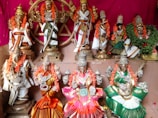 A collection of elaborately adorned Hindu deities is displayed on a tiered platform, each figure intricately decorated with vibrant garlands, jewelry, and colorful clothing. The background is a rich pink, complementing the overall aesthetic.