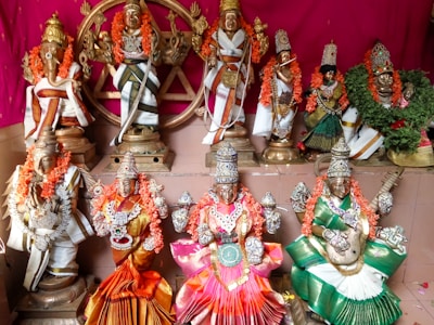A collection of elaborately adorned Hindu deities is displayed on a tiered platform, each figure intricately decorated with vibrant garlands, jewelry, and colorful clothing. The background is a rich pink, complementing the overall aesthetic.