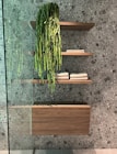 A modern bathroom interior featuring three wooden floating shelves mounted on a speckled grey stone wall. The top shelf holds a cascading green plant, while the middle and bottom shelves have neatly folded beige towels. A wooden cabinet is situated beneath the bottom shelf, with a glass panel visible on the left side.