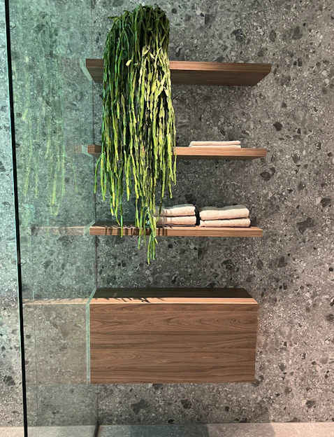A modern bathroom interior featuring three wooden floating shelves mounted on a speckled grey stone wall. The top shelf holds a cascading green plant, while the middle and bottom shelves have neatly folded beige towels. A wooden cabinet is situated beneath the bottom shelf, with a glass panel visible on the left side.