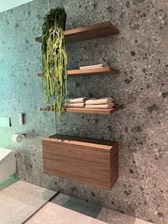 A modern bathroom interior with a speckled gray stone wall and a floating wooden cabinet. Three wooden shelves are mounted on the wall, two of them holding neatly folded towels and the top shelf featuring a cascading green plant. A toilet paper holder is attached to the wall, and a portion of a toilet is visible in the corner.