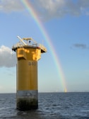 A towering yellow offshore structure stands in the ocean, labeled with 'B03,' with a vibrant rainbow arching across the blue sky. The structure is surrounded by calm waters reflecting light from the sun. In the distance, another similar structure is visible near the horizon.