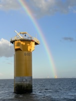 A towering yellow offshore structure stands in the ocean, labeled with 'B03,' with a vibrant rainbow arching across the blue sky. The structure is surrounded by calm waters reflecting light from the sun. In the distance, another similar structure is visible near the horizon.