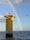 A towering yellow offshore structure stands in the ocean, labeled with 'B03,' with a vibrant rainbow arching across the blue sky. The structure is surrounded by calm waters reflecting light from the sun. In the distance, another similar structure is visible near the horizon.