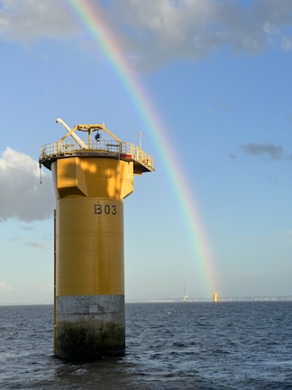 A towering yellow offshore structure stands in the ocean, labeled with 'B03,' with a vibrant rainbow arching across the blue sky. The structure is surrounded by calm waters reflecting light from the sun. In the distance, another similar structure is visible near the horizon.