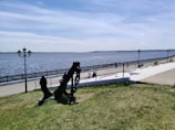 A large anchor is displayed on a grassy area near a walkway by the waterfront. The path is flanked by black ornate street lamps, and a metal fence runs along the edge of the water. The backdrop features a wide body of water under a clear blue sky with distant land visible on the horizon.