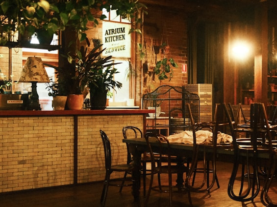 An indoor dining area with a cozy, rustic atmosphere. It features a mix of wooden and brick elements, with tables and chairs arranged in the space. Plants are placed throughout, contributing to a natural aesthetic, and there's a warm, dim lighting providing a calm ambiance.