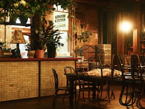 An indoor dining area with a cozy, rustic atmosphere. It features a mix of wooden and brick elements, with tables and chairs arranged in the space. Plants are placed throughout, contributing to a natural aesthetic, and there's a warm, dim lighting providing a calm ambiance.