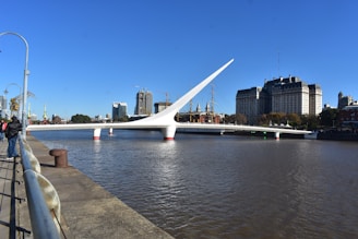 A modern white bridge with a striking, curved design spans over a wide river. In the background, a cityscape with high-rise buildings and a historic sailing ship is visible. The sky is clear and blue, creating a bright, sunny atmosphere. People are casually walking along the riverside, enjoying the view.