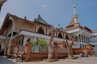 A historic mosque with a blend of traditional and modern architectural elements, featuring a large dome, a tall minaret, and intricate brickwork. The courtyard includes benches and pathways, and there are a couple of people in traditional attire.