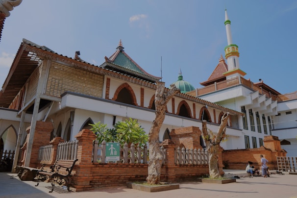 A historic mosque with a blend of traditional and modern architectural elements, featuring a large dome, a tall minaret, and intricate brickwork. The courtyard includes benches and pathways, and there are a couple of people in traditional attire.