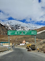 A mountainous landscape with a snow-capped peak in the background and a clear blue sky with some clouds. There is a large green directional road sign for various destinations including Baralacha La, Sarchu, Pang, Tanglang La, Upshi, and Leh, constructed by the Border Roads Organization. A road leads through the scene with construction equipment and structures such as buildings visible on the side.