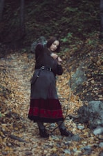 A person is standing in a forest path covered with fallen autumn leaves, dressed in medieval-style clothing. The individual appears to be holding a bow, suggesting a historical or fantasy theme. Trees and rocks surround the area, contributing to the rustic and natural setting.