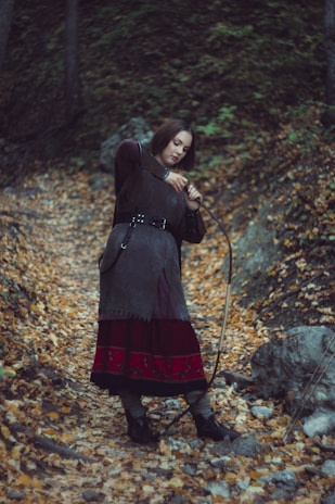 A person is standing in a forest path covered with fallen autumn leaves, dressed in medieval-style clothing. The individual appears to be holding a bow, suggesting a historical or fantasy theme. Trees and rocks surround the area, contributing to the rustic and natural setting.