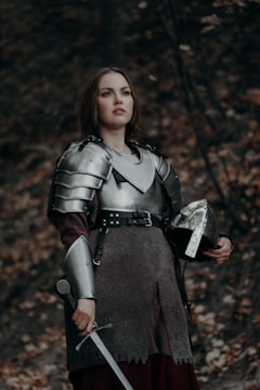 A person in medieval armor stands in a forest, holding a sword in one hand and a helmet in the other. The armor is shiny and metallic, contrasting with the earthy tones of the forest backdrop.