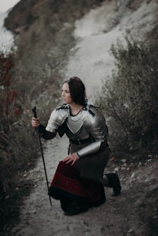A person in medieval armor is kneeling on a narrow dirt path in a natural setting. They hold a sword upright, resting it on the ground, surrounded by foliage and rocky terrain.