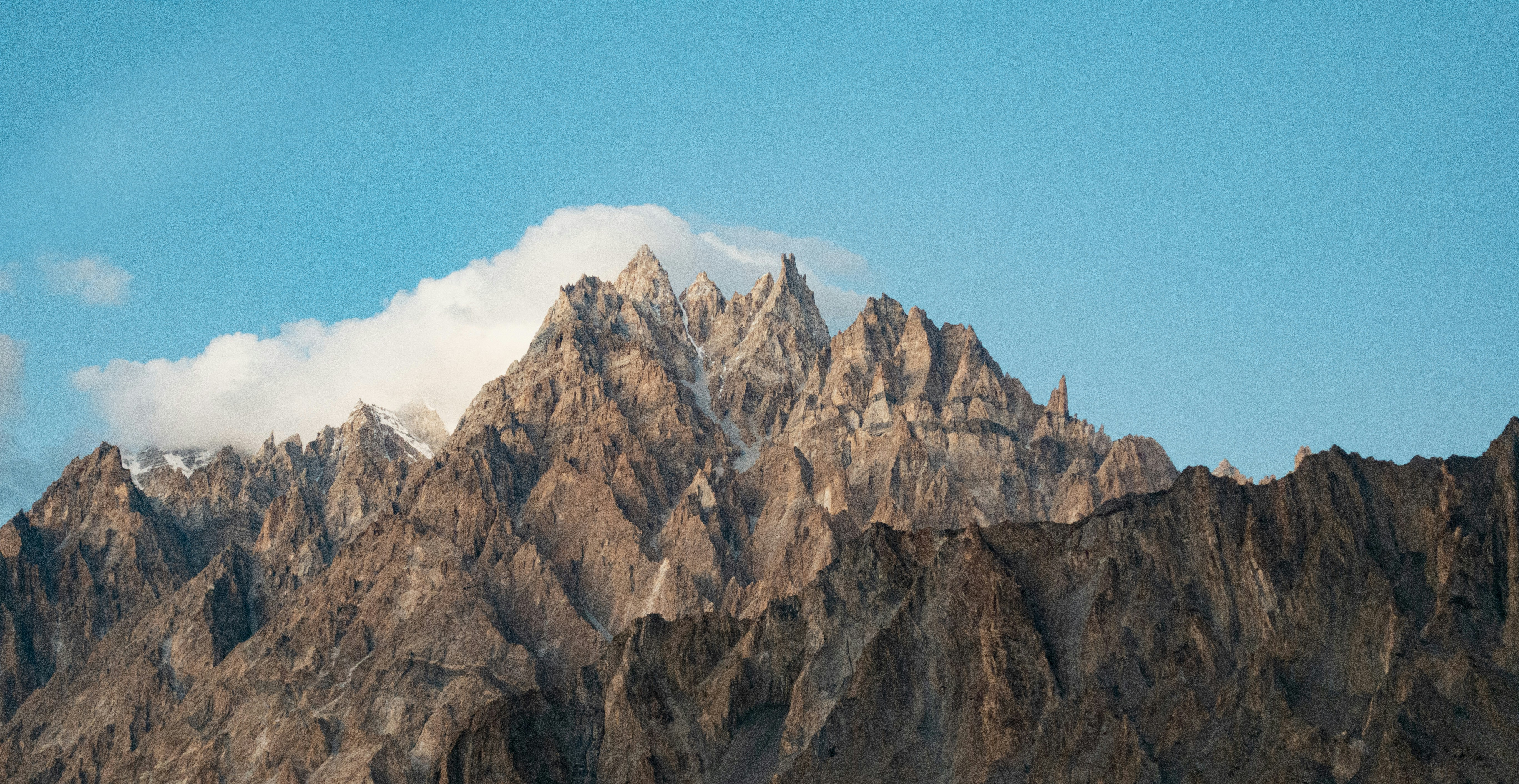 a very tall mountain with some clouds in the sky, A blue sky with sawtooth mountains and a cloud above them creates a picturesque and scenic landscape