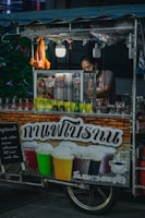 A street vendor, partially obscured, operates a small drink stall adorned with colorful beverage bottles. The stall's sign features images of iced drinks in various colors, suggesting offerings like iced coffee or tea. The vendor appears to prepare drinks amidst a dimly-lit outdoor environment with a canopy overhead and small hanging light fixtures.
