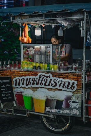 A street vendor, partially obscured, operates a small drink stall adorned with colorful beverage bottles. The stall's sign features images of iced drinks in various colors, suggesting offerings like iced coffee or tea. The vendor appears to prepare drinks amidst a dimly-lit outdoor environment with a canopy overhead and small hanging light fixtures.