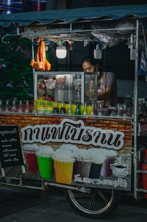 A street vendor, partially obscured, operates a small drink stall adorned with colorful beverage bottles. The stall's sign features images of iced drinks in various colors, suggesting offerings like iced coffee or tea. The vendor appears to prepare drinks amidst a dimly-lit outdoor environment with a canopy overhead and small hanging light fixtures.