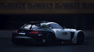 A sleek, high-performance race car with a prominent rear wing is captured in motion. The car features a black and white color scheme with various sponsorship logos visible on its body. The background shows a blurred view of a racetrack, and advertising banners for DeWalt are visible.