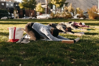Several people are practicing yoga outdoors on a grass field. They are all in a pose where they stretch forward with their arms extended and heads close to the ground. Personal items like a tote bag and shoes are placed nearby. The scene is set in a park with cars and trees in the background under clear skies.