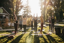 A group of people practicing yoga outdoors in a park setting, with their arms raised upwards. The scene is lit by a warm, golden sunlight filtering through tall trees, creating long shadows on the grass. A building, possibly a caf&eacute; or small shop, is visible in the background.