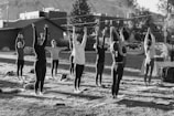 A group of people is practicing yoga outdoors on a grassy area, with their arms raised upwards. They are positioned on yoga mats and are surrounded by trees and buildings in the background, suggesting an urban park setting. The scene appears to be taking place during daylight, with shadows indicating the presence of sunlight.