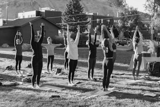 A group of people is practicing yoga outdoors on a grassy area, with their arms raised upwards. They are positioned on yoga mats and are surrounded by trees and buildings in the background, suggesting an urban park setting. The scene appears to be taking place during daylight, with shadows indicating the presence of sunlight.