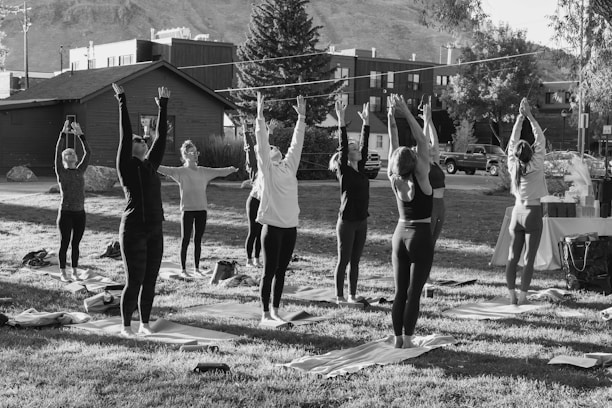 A group of people is practicing yoga outdoors on a grassy area, with their arms raised upwards. They are positioned on yoga mats and are surrounded by trees and buildings in the background, suggesting an urban park setting. The scene appears to be taking place during daylight, with shadows indicating the presence of sunlight.