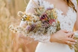 A bride holding a bouquet of dried flowers. The bride wears a lace wedding dress with intricate patterns. The bouquet features a mix of dried blooms in earthy tones, including purples, whites, and greens.