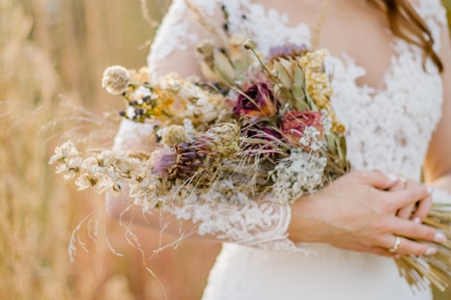 A bride holding a bouquet of dried flowers. The bride wears a lace wedding dress with intricate patterns. The bouquet features a mix of dried blooms in earthy tones, including purples, whites, and greens.