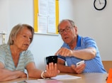 An older woman and man are sitting at a table in a well-lit room. The man, wearing glasses and a blue checkered shirt, is pointing at a smartphone held by the woman. They appear to be engaged in a discussion, possibly explaining or learning something about the device. In the background, there is a clock on the wall and some documents pinned up.