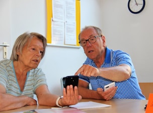 An older woman and man are sitting at a table in a well-lit room. The man, wearing glasses and a blue checkered shirt, is pointing at a smartphone held by the woman. They appear to be engaged in a discussion, possibly explaining or learning something about the device. In the background, there is a clock on the wall and some documents pinned up.