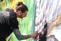 A person with long hair tied back is spray painting a colorful mural on a wall. Their arm, covered in tattoos, is extended as they hold a spray paint can. The mural features a blend of greens, purples, and browns, creating a dynamic and artistic scene.