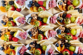 A collection of small paper trays filled with an assortment of snacks including fresh strawberries, raspberries, blueberries, blackberries, green grapes, almonds, dried apricots, crackers, slices of cheese, and rolled salami. The arrangement is colorful and appealing, presenting a variety of textures and flavors.