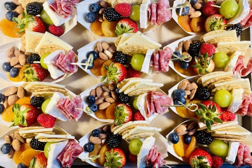 A collection of small paper trays filled with an assortment of snacks including fresh strawberries, raspberries, blueberries, blackberries, green grapes, almonds, dried apricots, crackers, slices of cheese, and rolled salami. The arrangement is colorful and appealing, presenting a variety of textures and flavors.