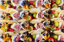 A collection of small paper trays filled with an assortment of snacks including fresh strawberries, raspberries, blueberries, blackberries, green grapes, almonds, dried apricots, crackers, slices of cheese, and rolled salami. The arrangement is colorful and appealing, presenting a variety of textures and flavors.