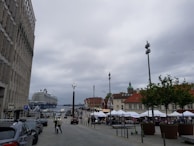 A large cruise ship is docked at a port near a street lined with buildings and market stalls. People are walking around, and several cars are parked along the street. The sky is overcast, and there are pigeons visible in the sky. Trees and lamp posts are also present along the street.