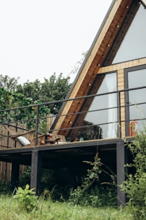 An A-frame house with large glass windows and a wooden exterior is situated on a deck surrounded by lush greenery. The area is characterized by a simple yet cozy setting, with patio furniture that includes a chair and a small table with decorative items.