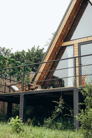 An A-frame house with large glass windows and a wooden exterior is situated on a deck surrounded by lush greenery. The area is characterized by a simple yet cozy setting, with patio furniture that includes a chair and a small table with decorative items.