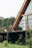 An A-frame house with large glass windows and a wooden exterior is situated on a deck surrounded by lush greenery. The area is characterized by a simple yet cozy setting, with patio furniture that includes a chair and a small table with decorative items.