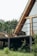 An A-frame house with large glass windows and a wooden exterior is situated on a deck surrounded by lush greenery. The area is characterized by a simple yet cozy setting, with patio furniture that includes a chair and a small table with decorative items.