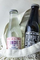 Two glass bottles of Indi & Co organic beverages are placed side by side in a cream-colored textile basket. One bottle appears clear with a strawberry label, while the other is dark with a kola soda label.