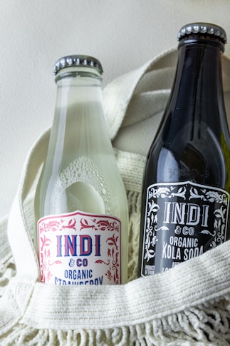 Two glass bottles of Indi & Co organic beverages are placed side by side in a cream-colored textile basket. One bottle appears clear with a strawberry label, while the other is dark with a kola soda label.
