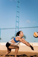 A person is playing beach volleyball, preparing to receive or bump the ball. The individual is wearing a sleeveless top and shorts, with a volleyball flying toward them. The background includes a blue sky and a net above a sand court.